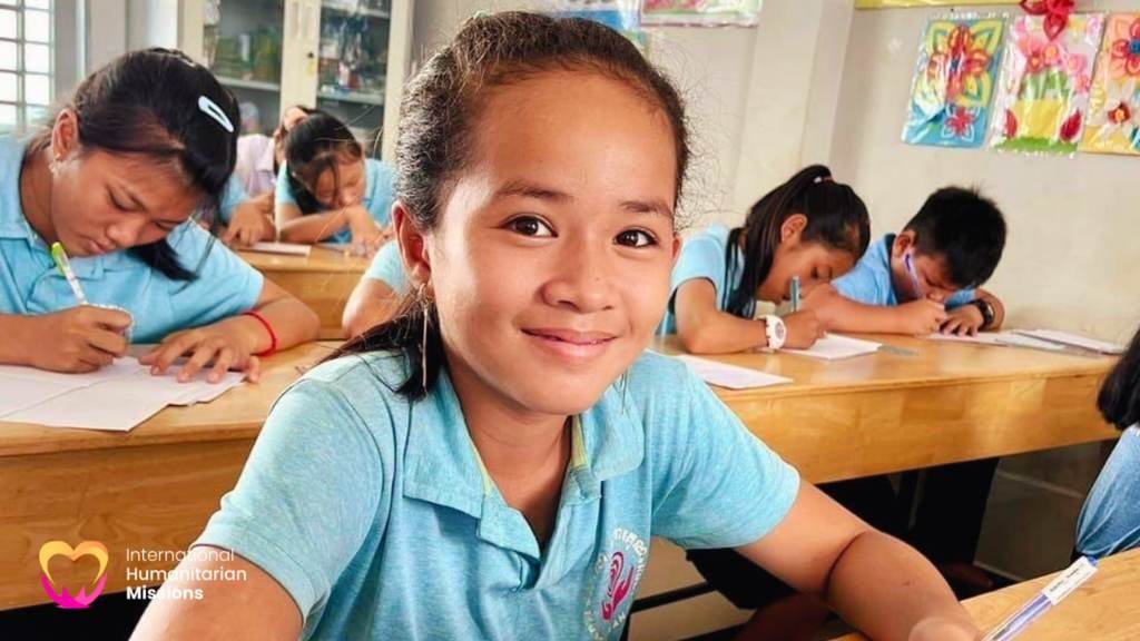 Tao, smiling at the camera while sitting at her classroom desk with classmates writing in the background